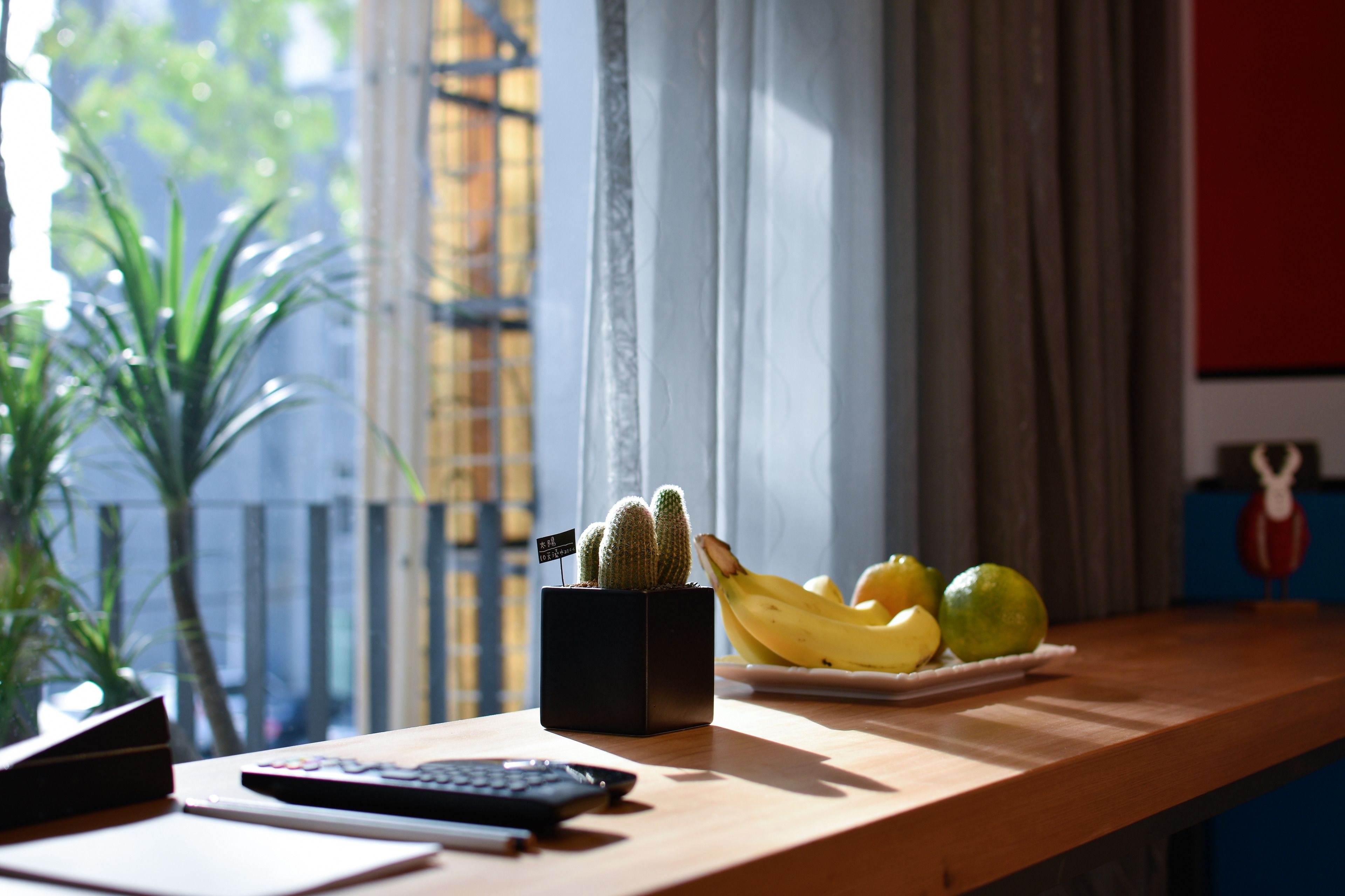 A wooden desk with a black cactus planter, a plate of bananas and limes, and a remote control.