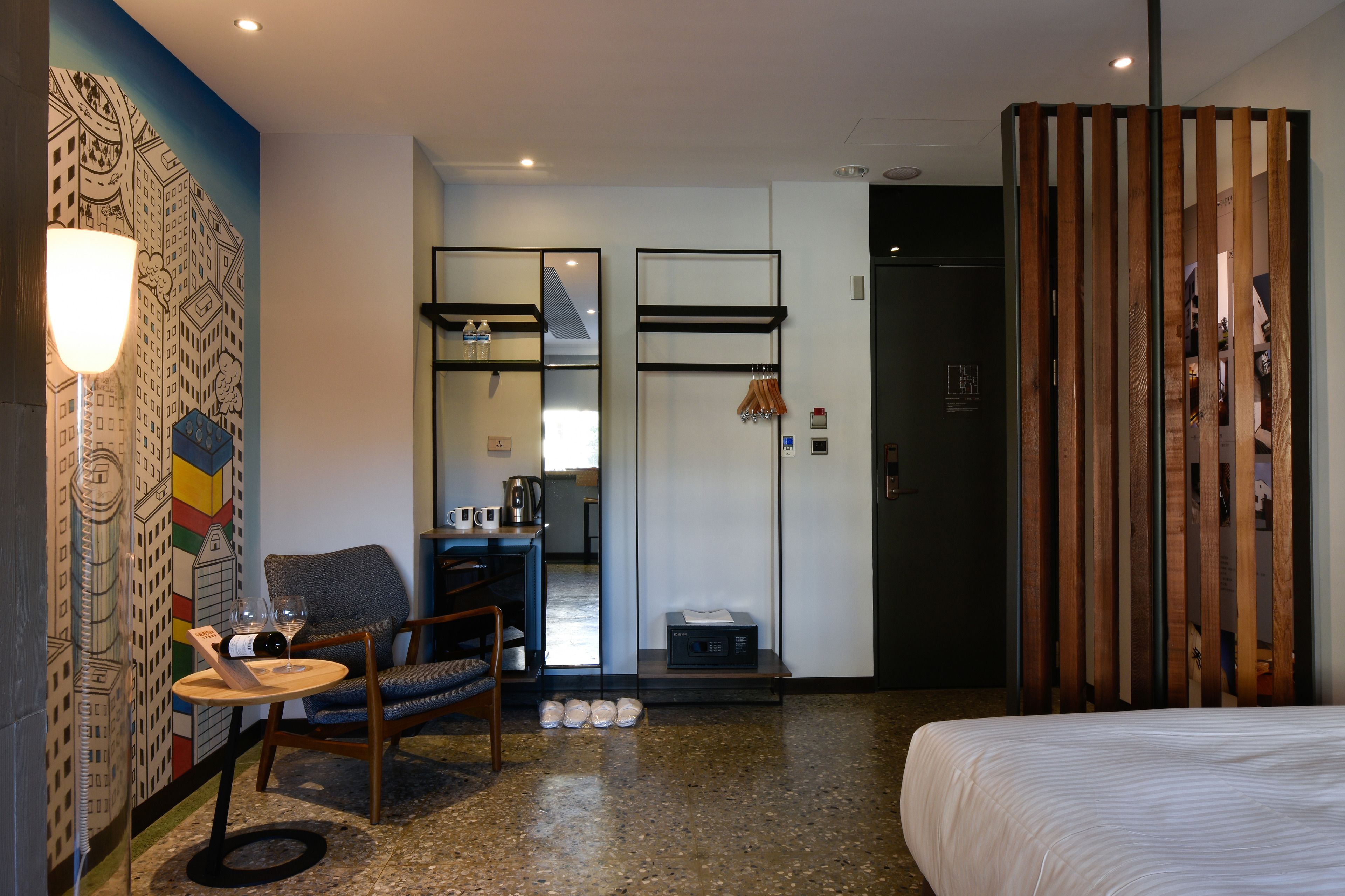 A hotel room with a bed, a chair, a small table with a wine bottle and glasses, a coffee machine, and a mirror.