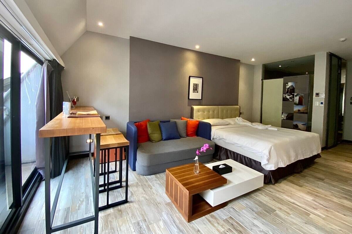 A modern hotel room with a bed, sofa, wooden coffee table, and a desk with a chair.