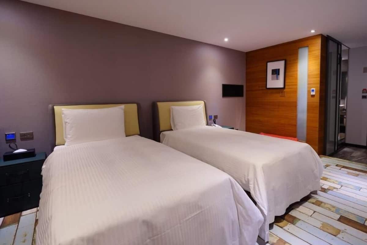 A hotel room with two beds, a wooden accent wall, a small desk with a phone, and a glass door leading to a bathroom.