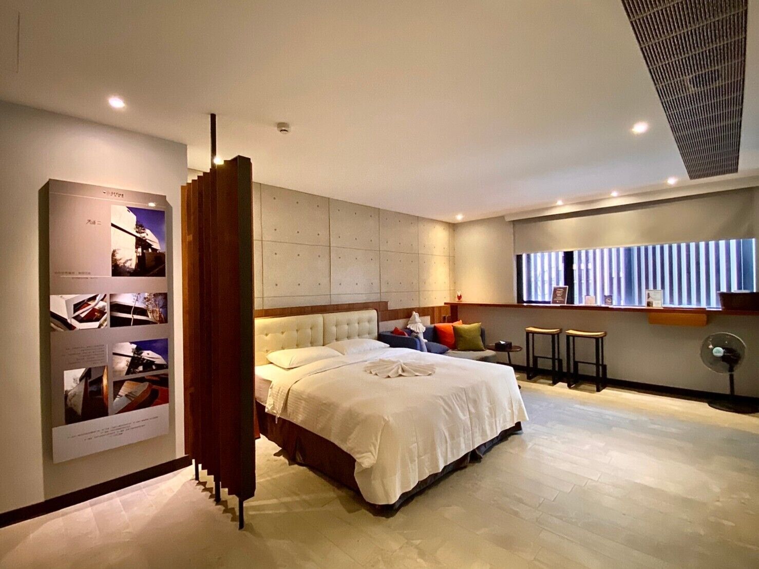 Una habitación de hotel moderna con una cama grande, una zona de estar con taburetes, una mesita y una ventana con persianas.