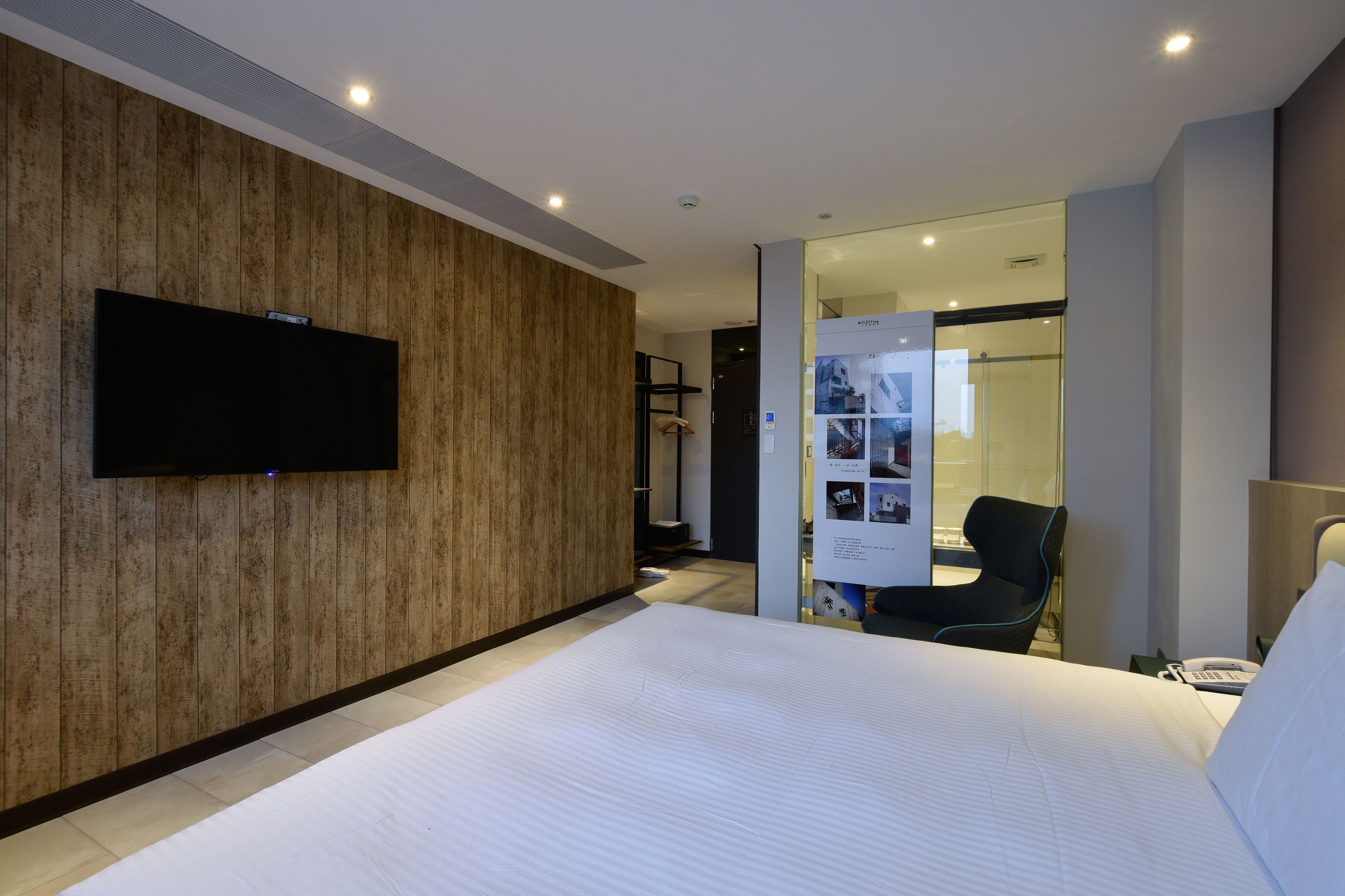 A modern hotel room with a large bed, a flat-screen TV mounted on a wooden wall, a glass shower enclosure, and a seating area with a chair and a small table.