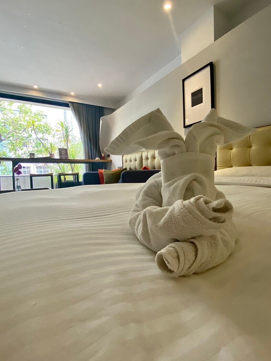 A neatly made bed with a large, rolled-up towel in the center, a tufted headboard, and a framed picture on the wall.