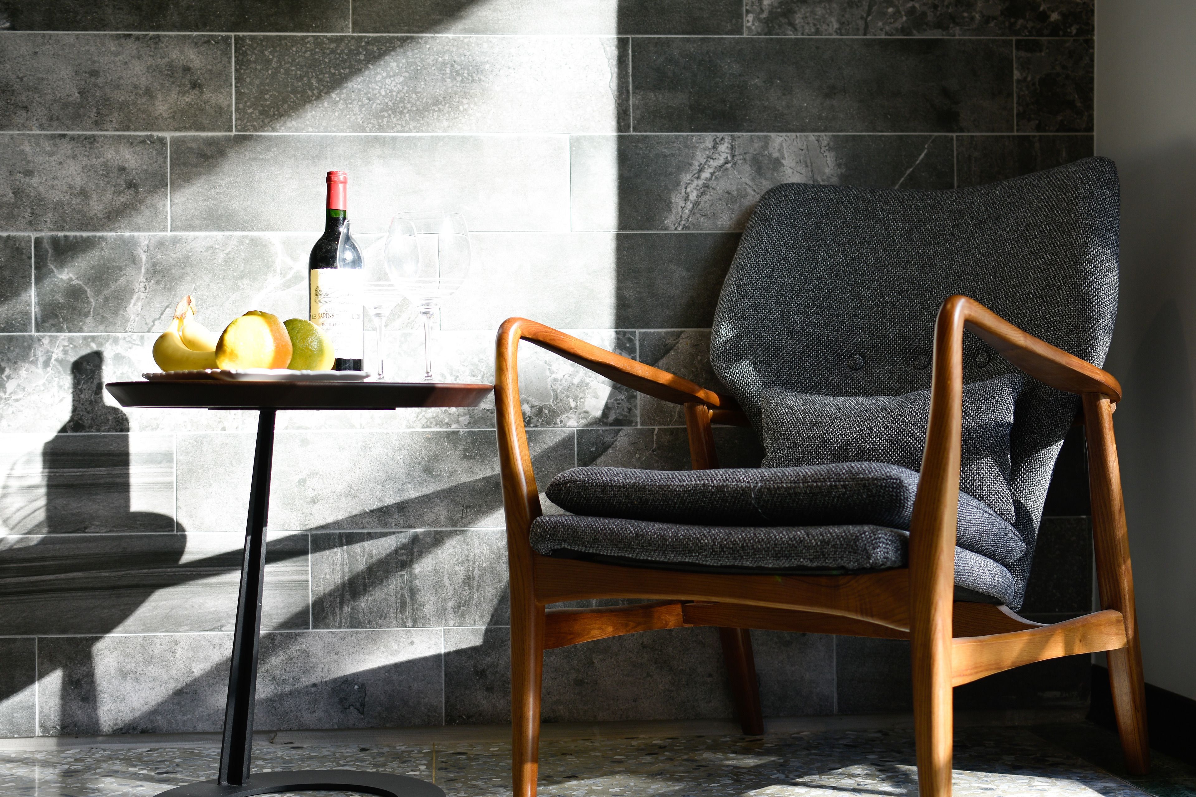 A modern chair with a wooden frame and grey upholstered seat, positioned next to a table with a bottle of wine, a glass, and some fruit.