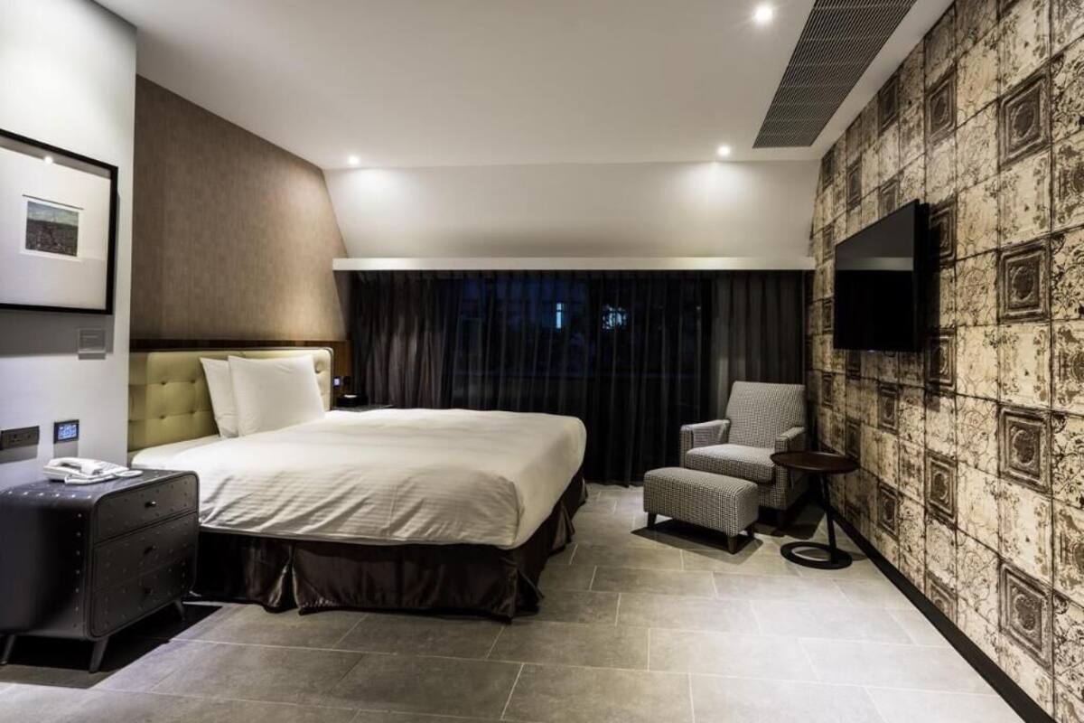 A modern hotel room with a large bed, a flat-screen TV, a seating area, and a wall-mounted TV.