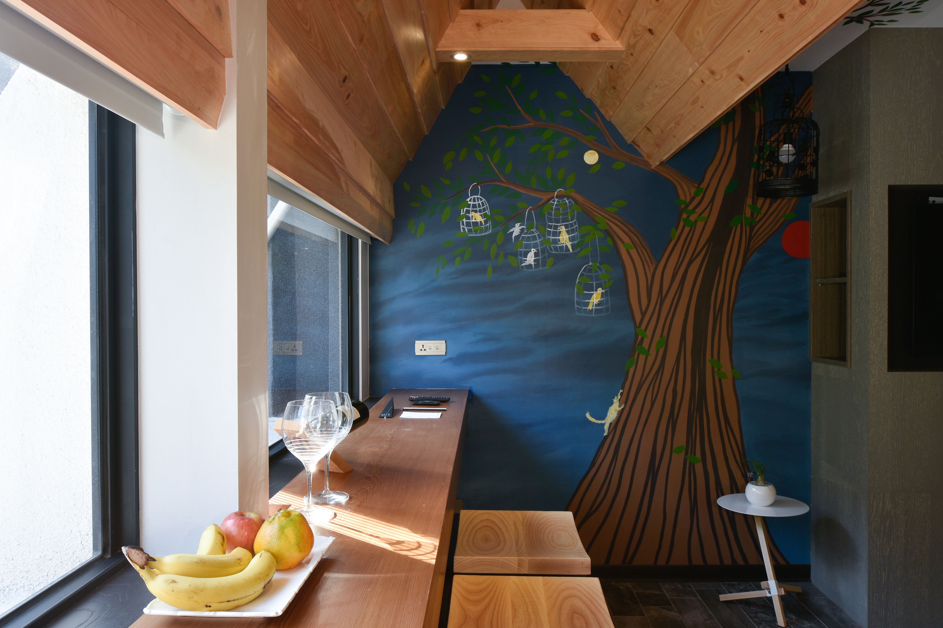 Une pièce avec une table en bois, une assiette de fruits, un verre et une fresque murale représentant un arbre avec des cages à oiseaux.