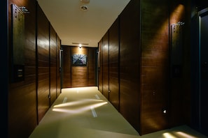 Un estrecho pasillo con paneles de madera y puertas numeradas, que conduce a una habitación con un gran ventanal y un cuadro.