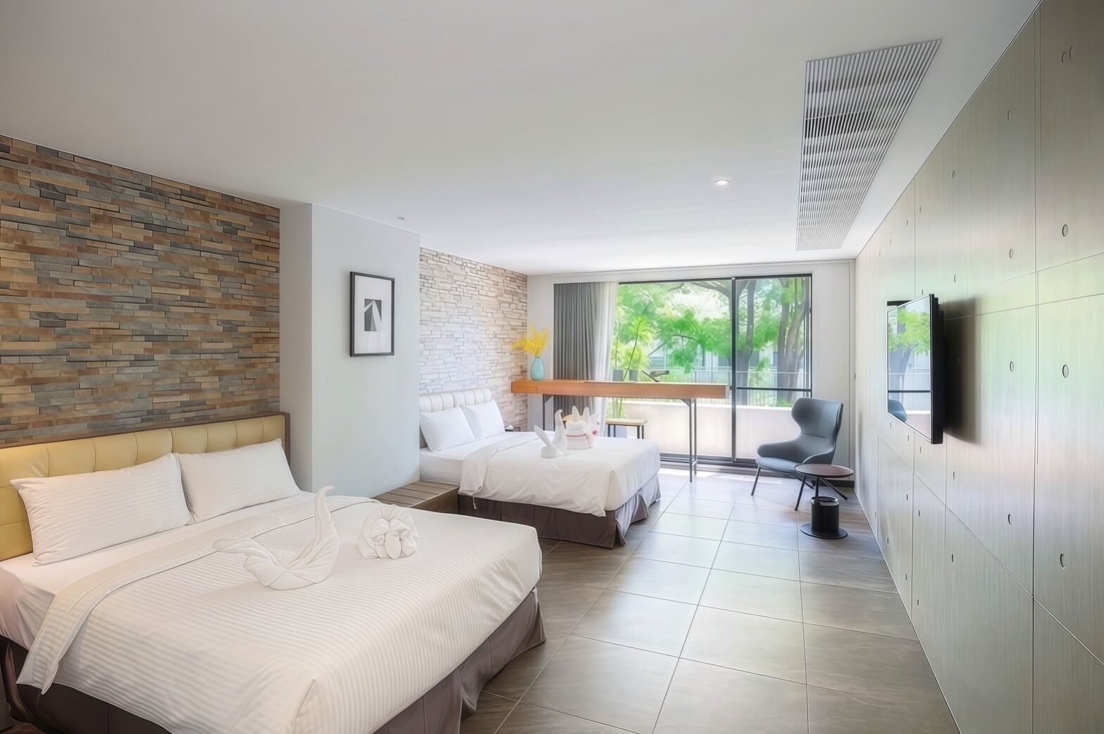 A hotel room with two beds, a desk, a chair, a TV, and a large window with a view of greenery.