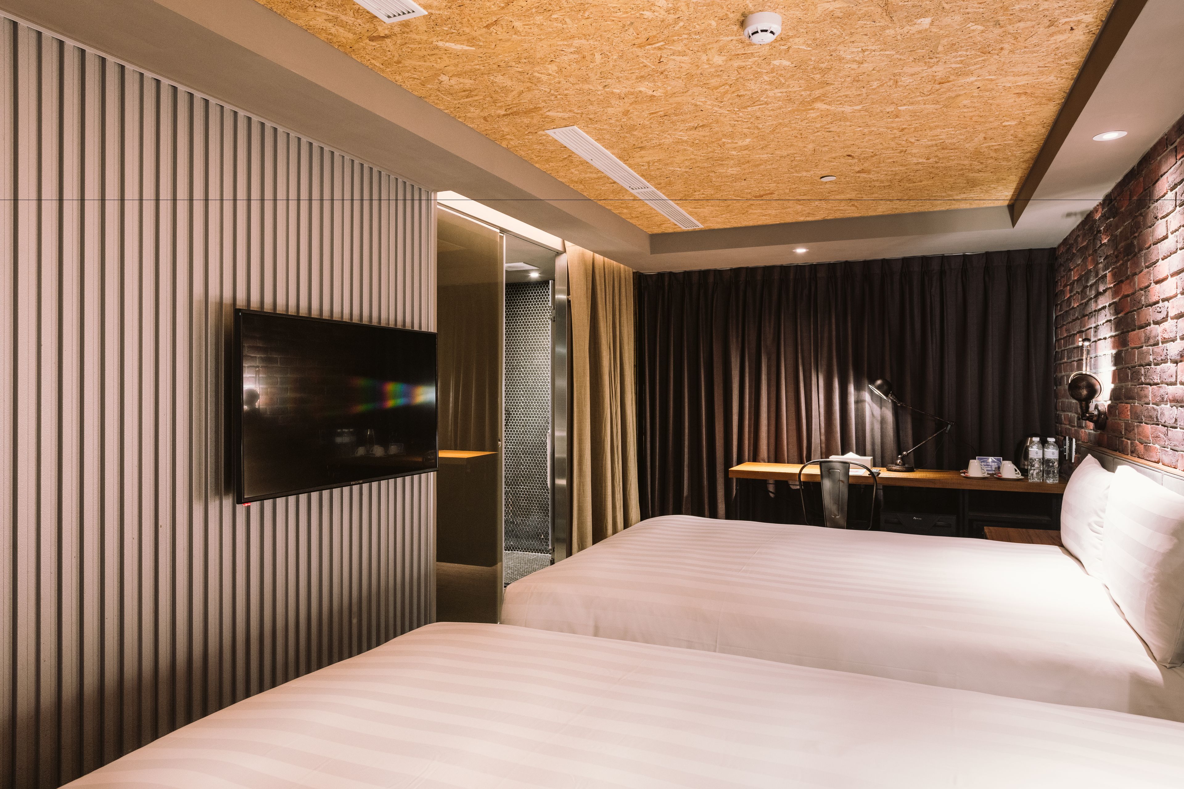 A modern hotel room with two beds, a flat-screen TV mounted on the wall, a desk with a lamp, and a brick accent wall.