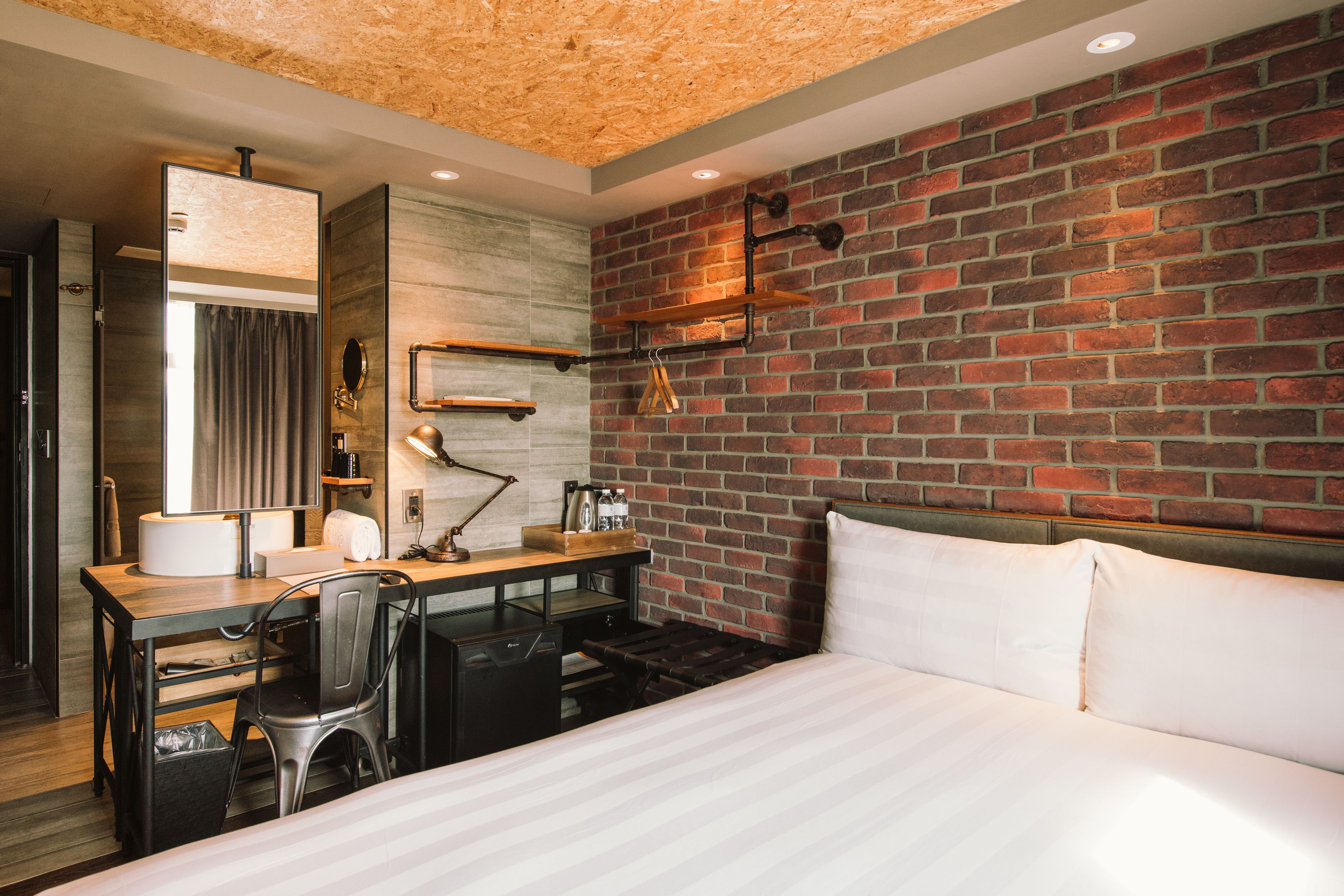 A hotel room with a brick wall, a wooden desk with a chair, a bed with white linens, and a modern bathroom with a mirror and towel rack.
