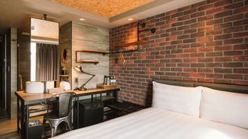 A hotel room with a brick wall, a wooden desk with a chair, a bed with white linens, and a modern bathroom with a mirror and towel rack.