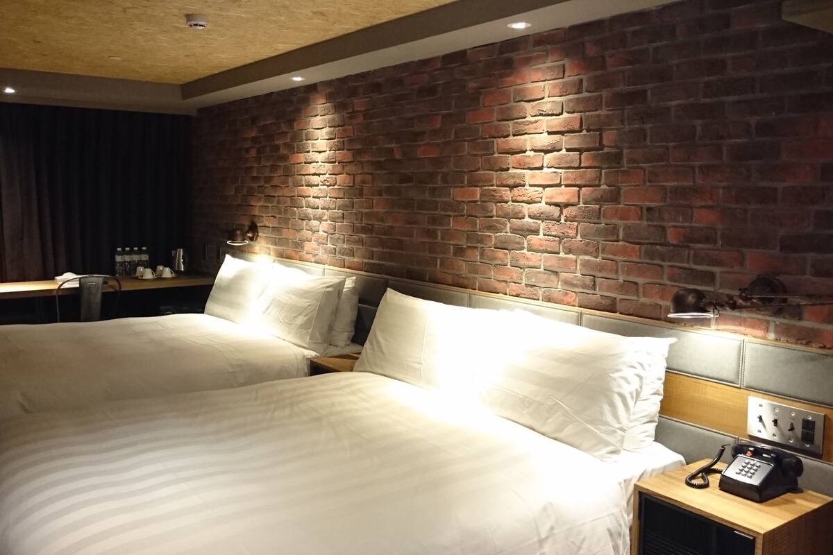 Ein Hotelzimmer mit zwei Betten, einer Ziegelwand, einem Schreibtisch mit Stuhl und einem Telefon auf einem Nachttisch aus Holz.