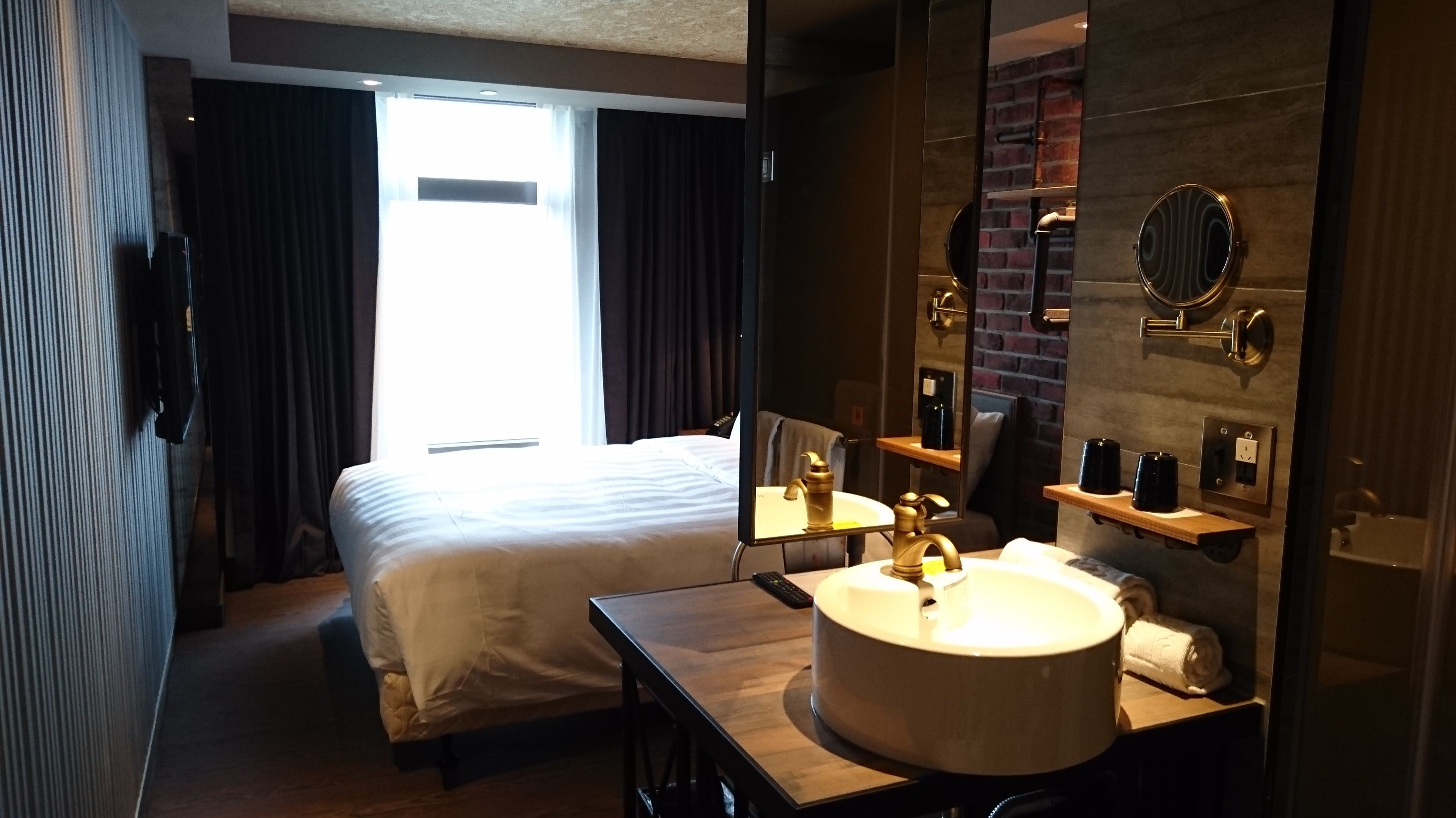 Quarto de hotel com cama, mesa de cabeceira, espelho redondo, pia e vista para o exterior pela janela.