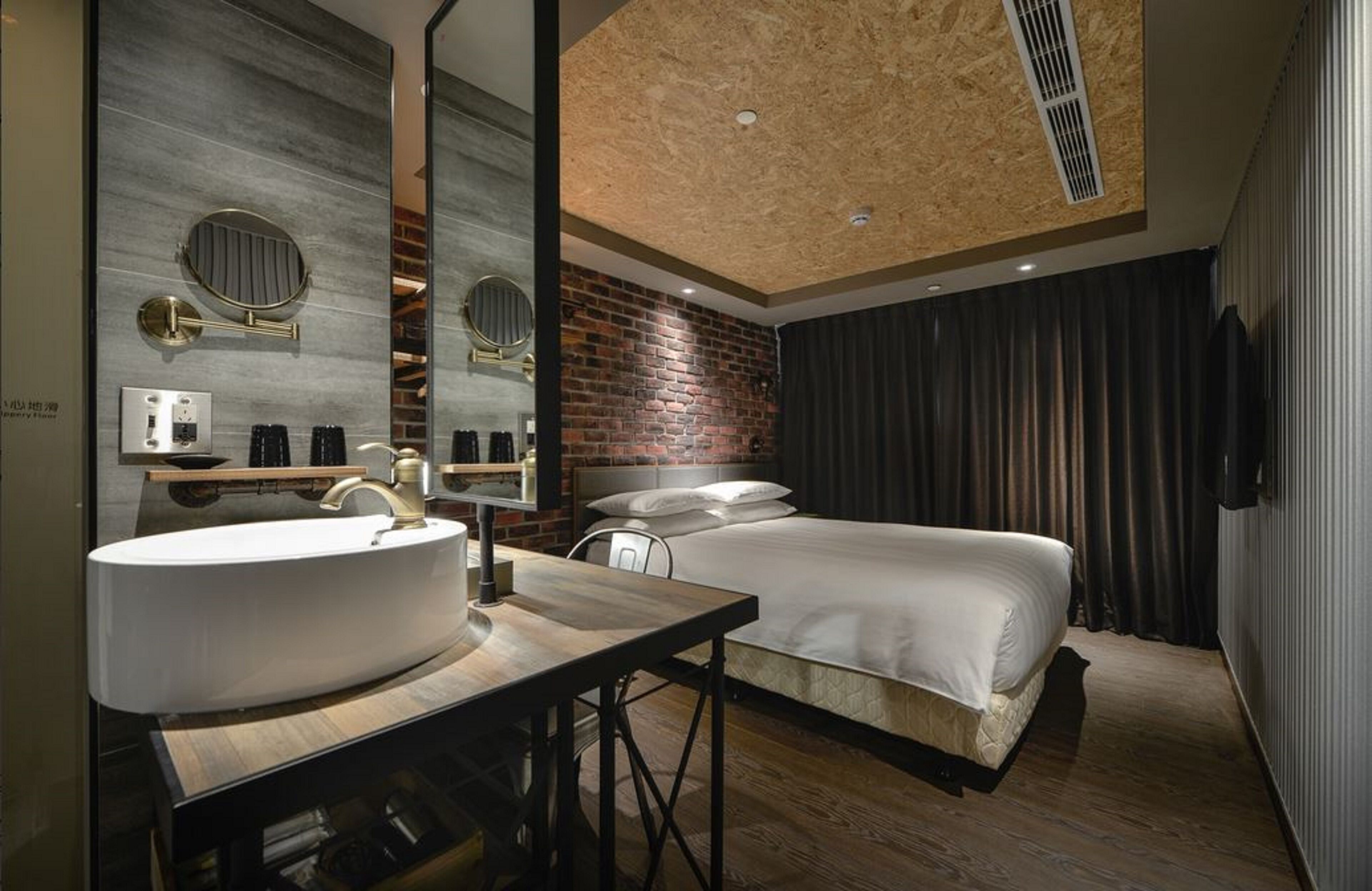 A modern hotel room with a large round mirror, a wooden vanity, a bed with white linens, a brick accent wall, and a ceiling with a wooden panel design.