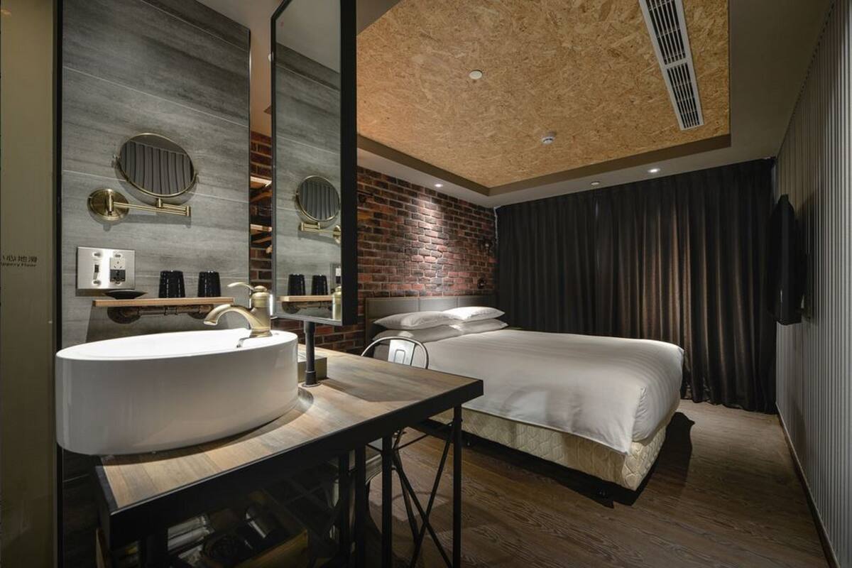 Ein modernes Hotelzimmer mit einem großen, runden Spiegel, einem hölzernen Schminktisch, einem Bett mit weißen Leinen, einer Wand mit Ziegeloptik und einer Decke mit Holzpaneelen.