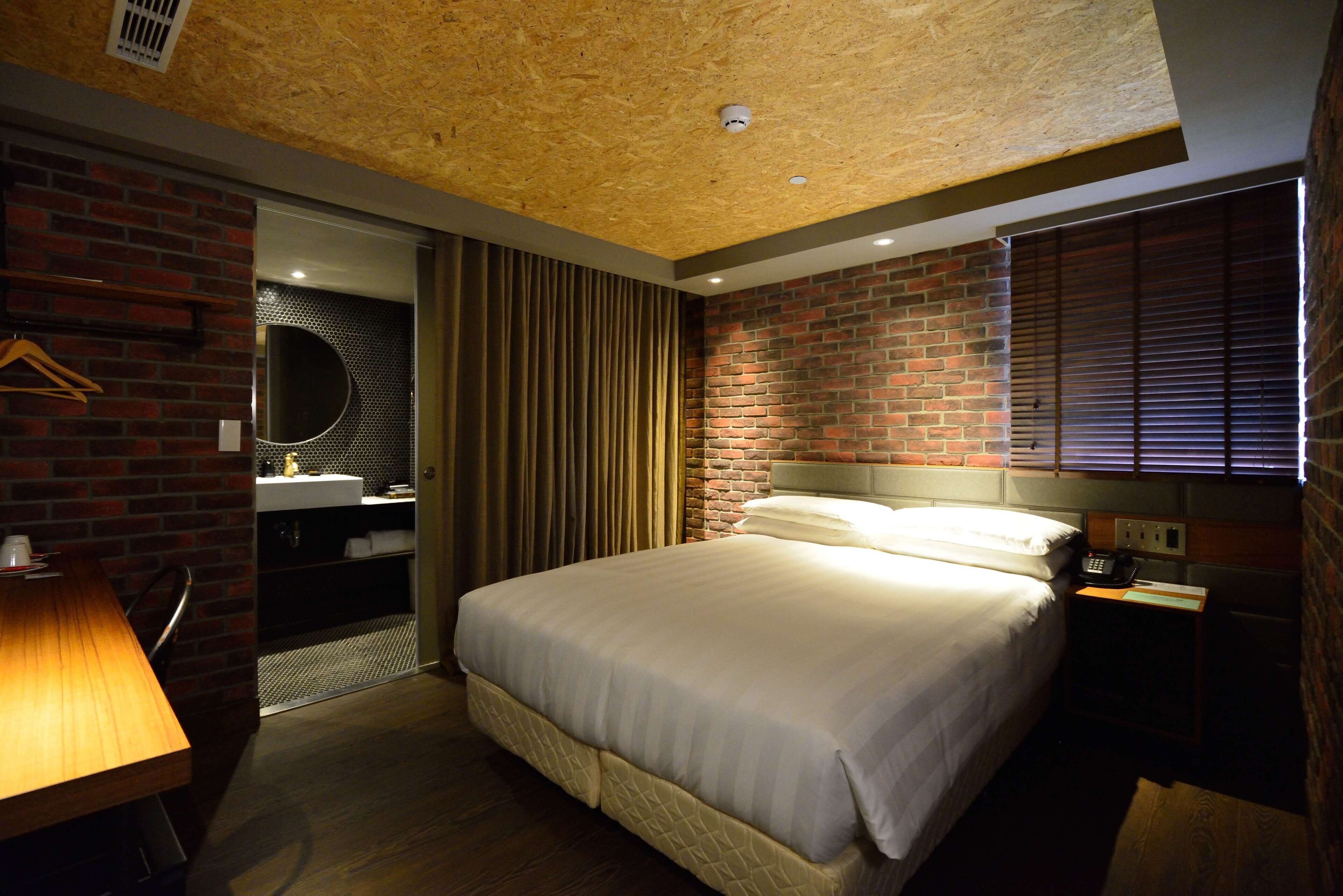 A hotel room with a brick wall, a bed with white bedding, a desk with a lamp, and a bathroom visible through a door.
