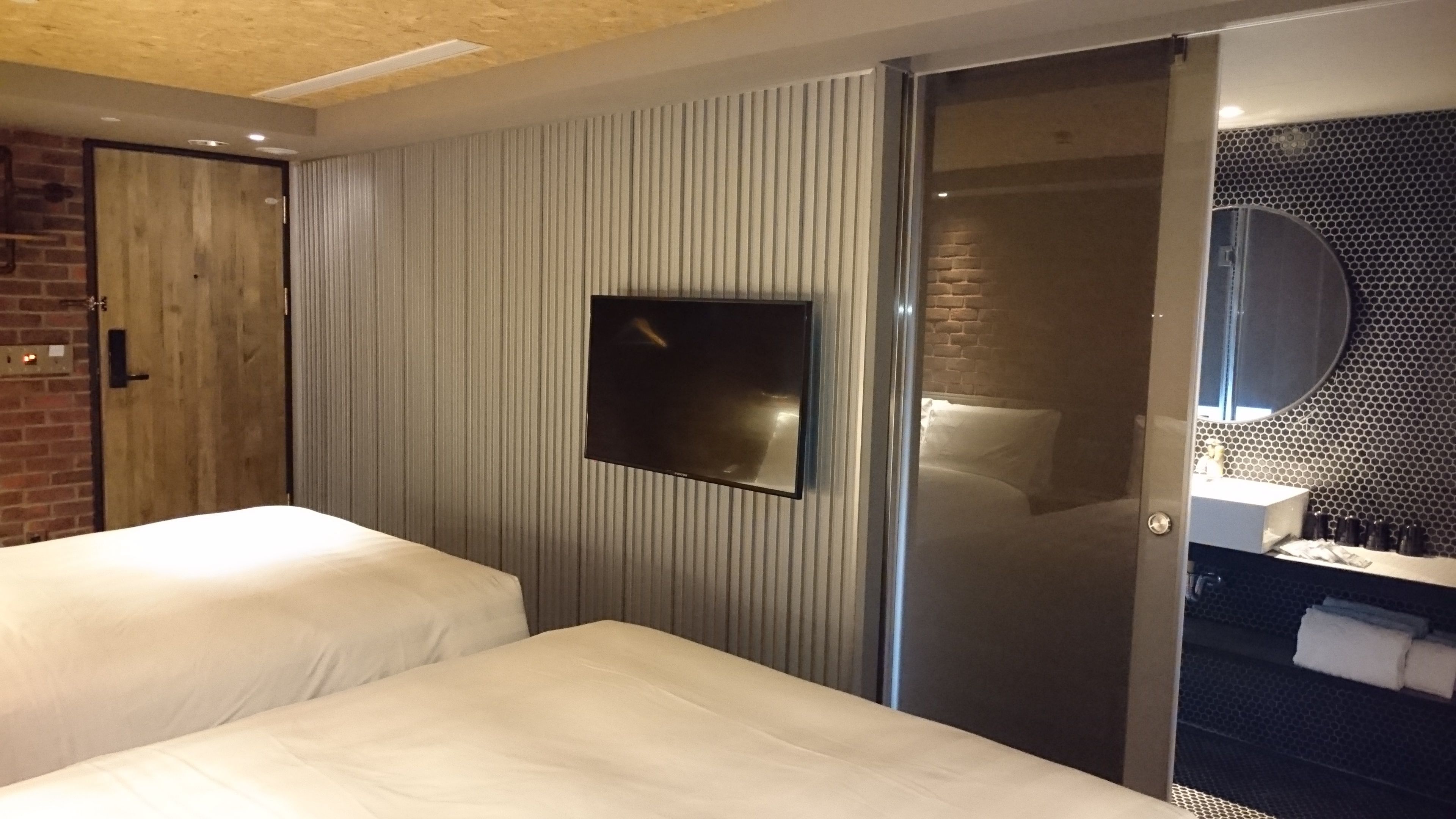 Une chambre d’hôtel avec deux lits, un téléviseur fixé au mur, une porte coulissante donnant accès à une salle de bain et un mur en briques.