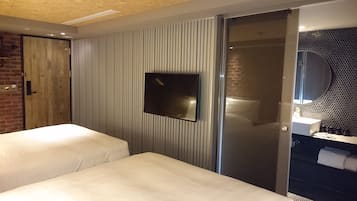 Une chambre d’hôtel avec deux lits, un téléviseur fixé au mur, une porte coulissante donnant accès à une salle de bain et un mur en briques.