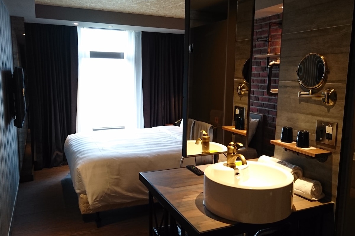 A hotel room with a bed, a bedside table with a lamp, a large mirror, a sink, and a view of the outside.