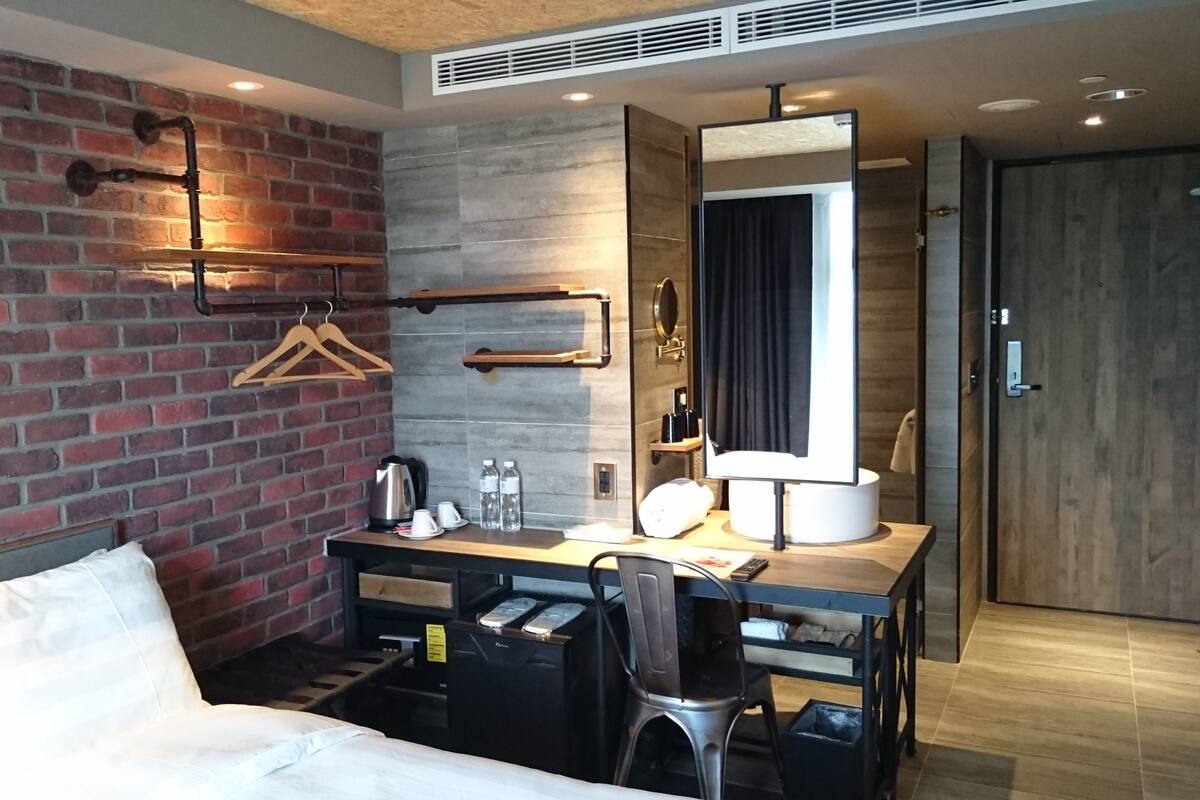 Ein Hotelzimmer mit Backsteinwand, Holztisch, Spiegel, Bett und Tür.