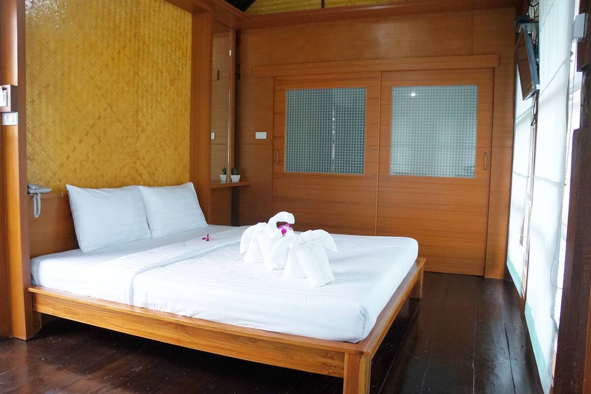 Una cama bien hecha con ropa de cama blanca y un arreglo de toallas, un cabecero de madera, una mesita de noche pequeña con una lámpara, y una ventana con diseño de cuadrícula.