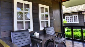 A wooden porch with two wooden chairs and a table, a window with a white frame, and a view of a house with a tiled roof. - Chanpraya Resort (Chanthaburi)
