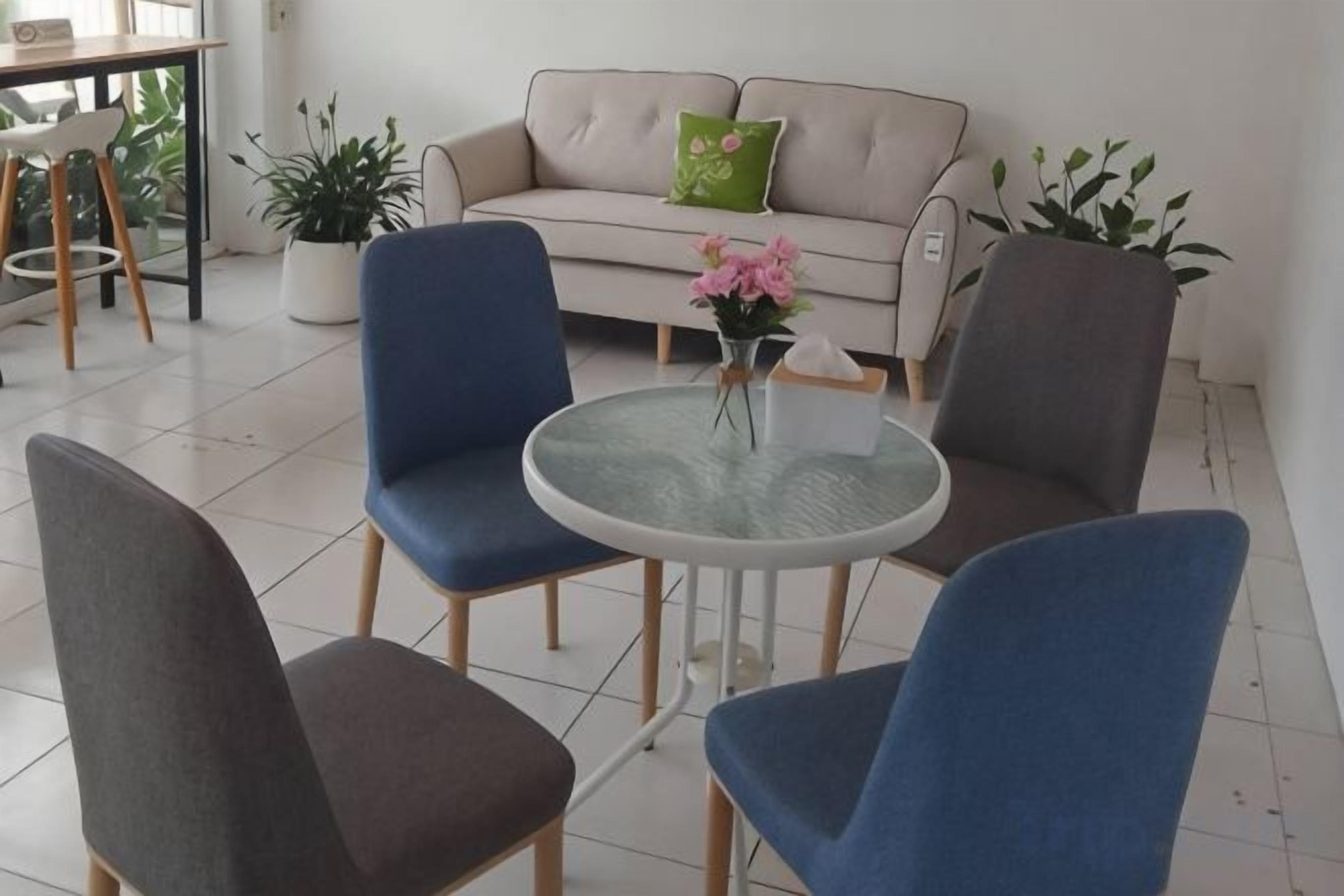 A room with a round glass table surrounded by four chairs, a sofa with a green pillow, and potted plants.