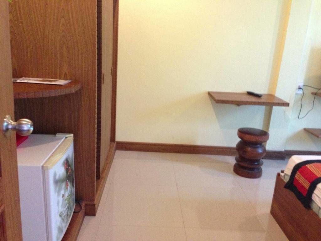 A room with a wooden cabinet, a small wooden stool, a white refrigerator, and a wooden shelf on the wall.