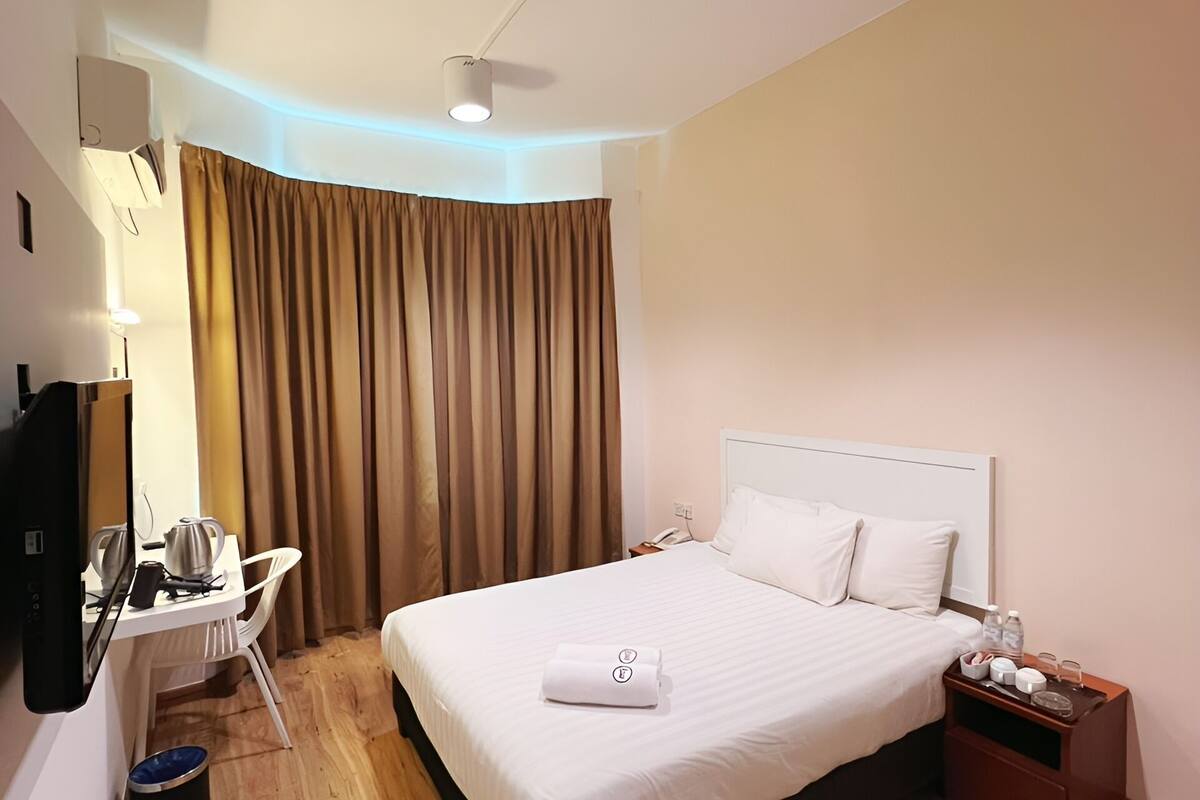 A hotel room with a neatly made bed, a desk with a chair, a bedside table with a lamp, and a ceiling light.