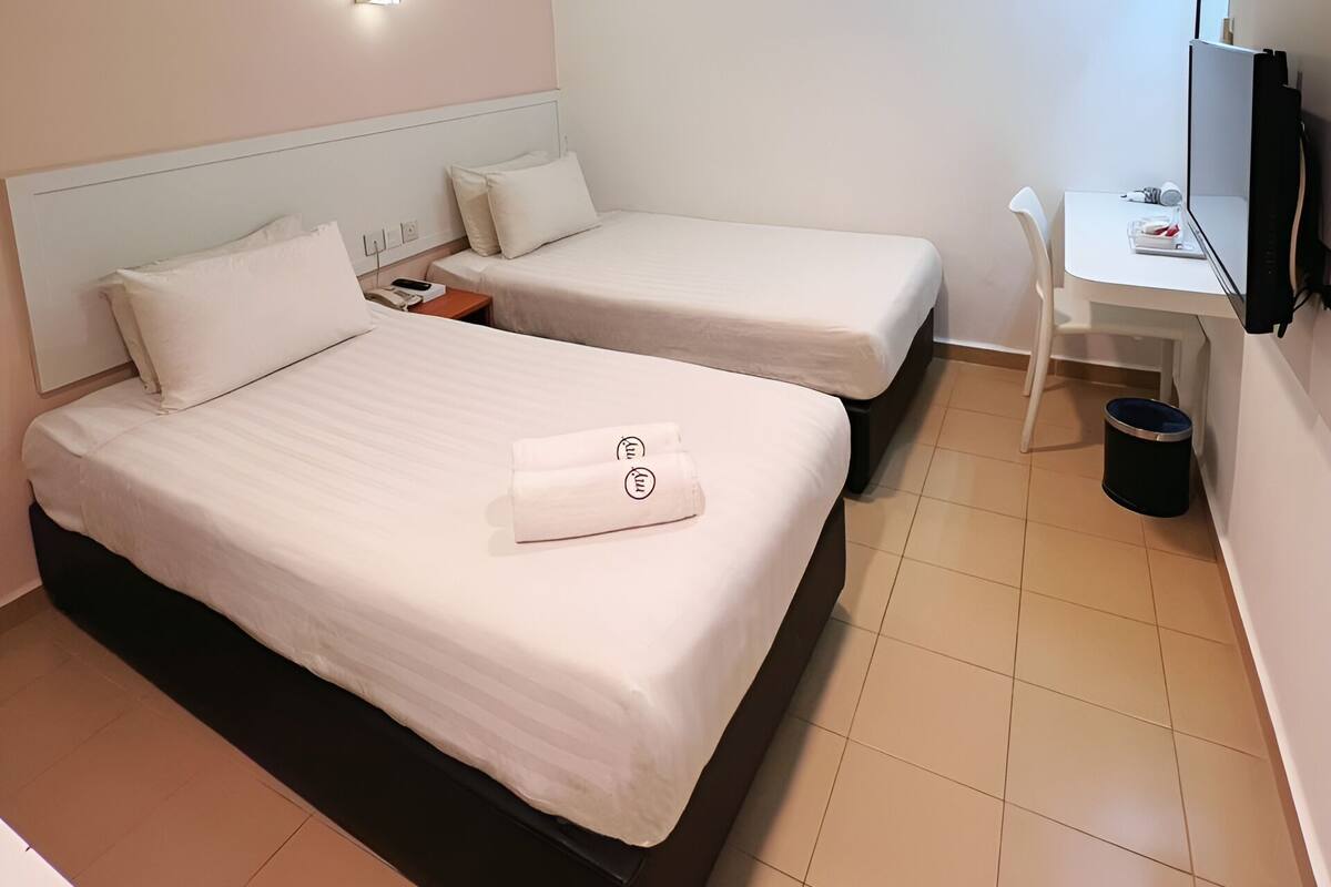 A hotel room with two beds, a desk, a chair, a TV, and a trash bin.
