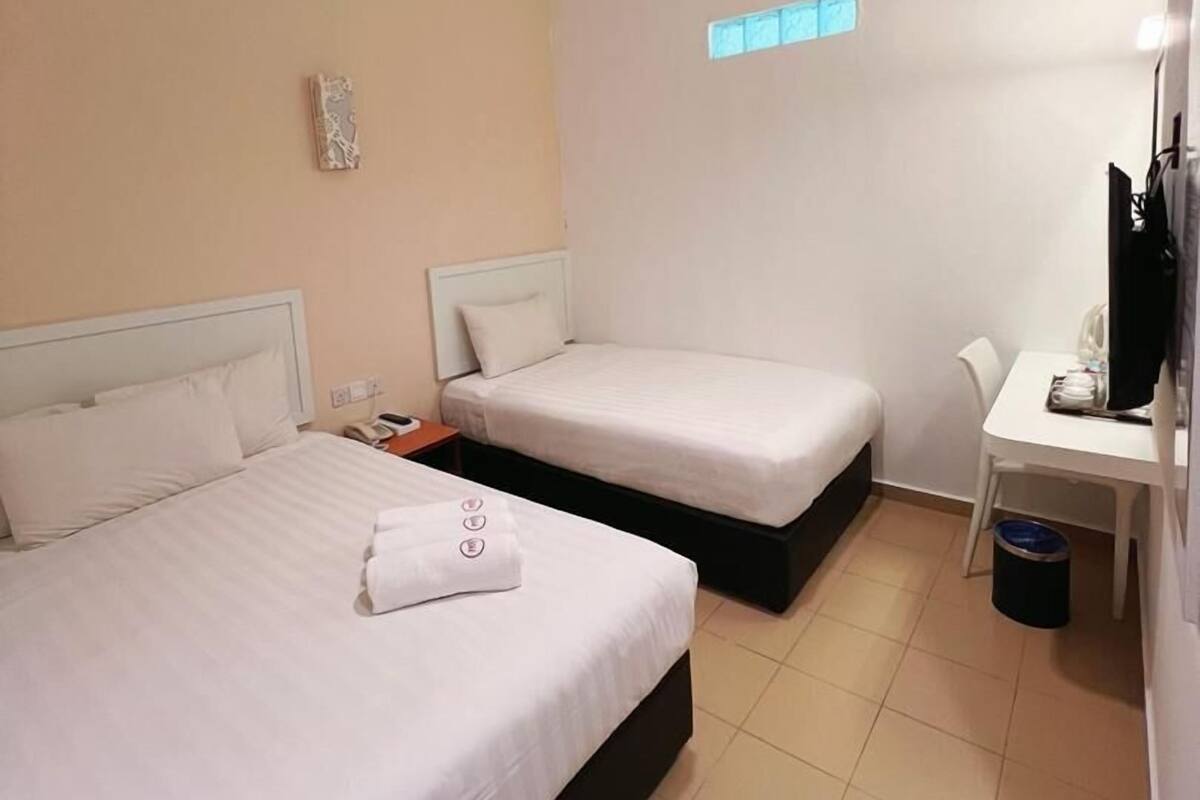 A hotel room with two beds, a desk, a chair, a small window, and a ceiling light.