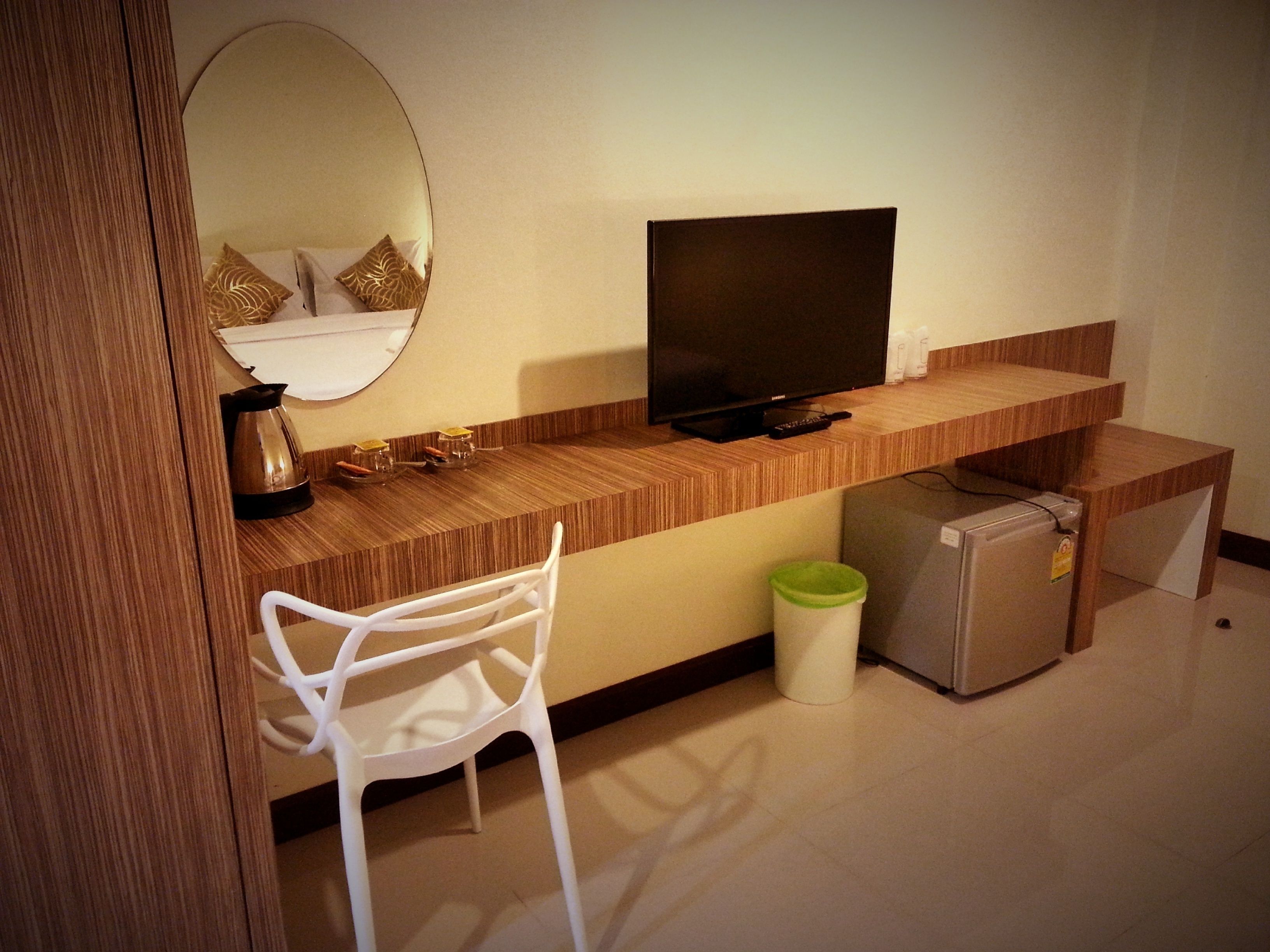 A compact hotel room with a wooden desk, a television, a white chair, a small refrigerator, and a trash bin.