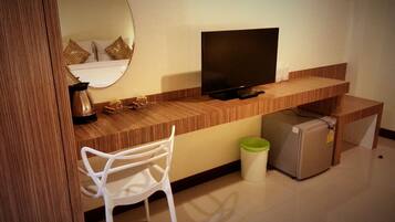 A compact hotel room with a wooden desk, a television, a white chair, a small refrigerator, and a trash bin.