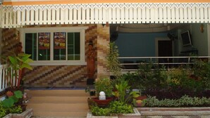 Un edificio con fachada de tejas, un toldo decorativo en la entrada y un pequeño jardín con plantas variadas y una fuente de agua.