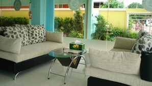 Un moderno hall de hotel con un sofá y una mesa de centro de vidrio, una recepción y vistas al área exterior con plantas y un edificio amarillo.