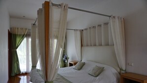 A four-poster bed with a wooden frame and white bedding, a wooden nightstand with a microwave, and a green curtain on the window. - Ban Rim Nam Riverside Resort (Nan)