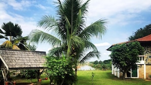 A tropical garden with a palm tree, a brick structure, and a building with a tiled roof. - Ban Rim Nam Riverside Resort (Nan)