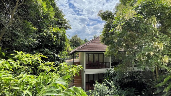 A two-story house surrounded by dense greenery, with a balcony on the upper floor.