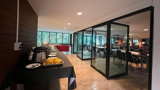 A modern dining area with a long table set for a meal, a buffet with various dishes, and a glass partition separating the dining area from another room. - Green Hill Resort (Pua)