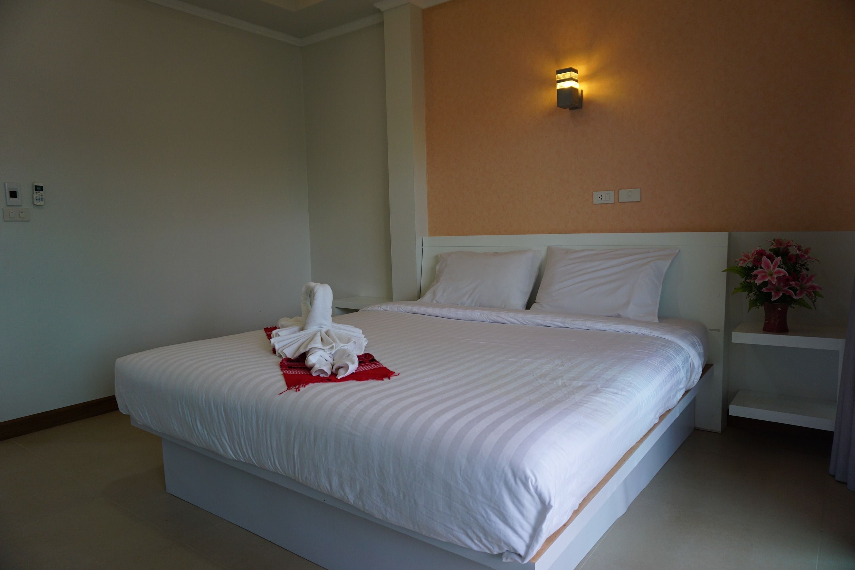 Una cama bien tendida con sábanas blancas y una toalla, un aplique de pared y un estante con un jarrón de flores.