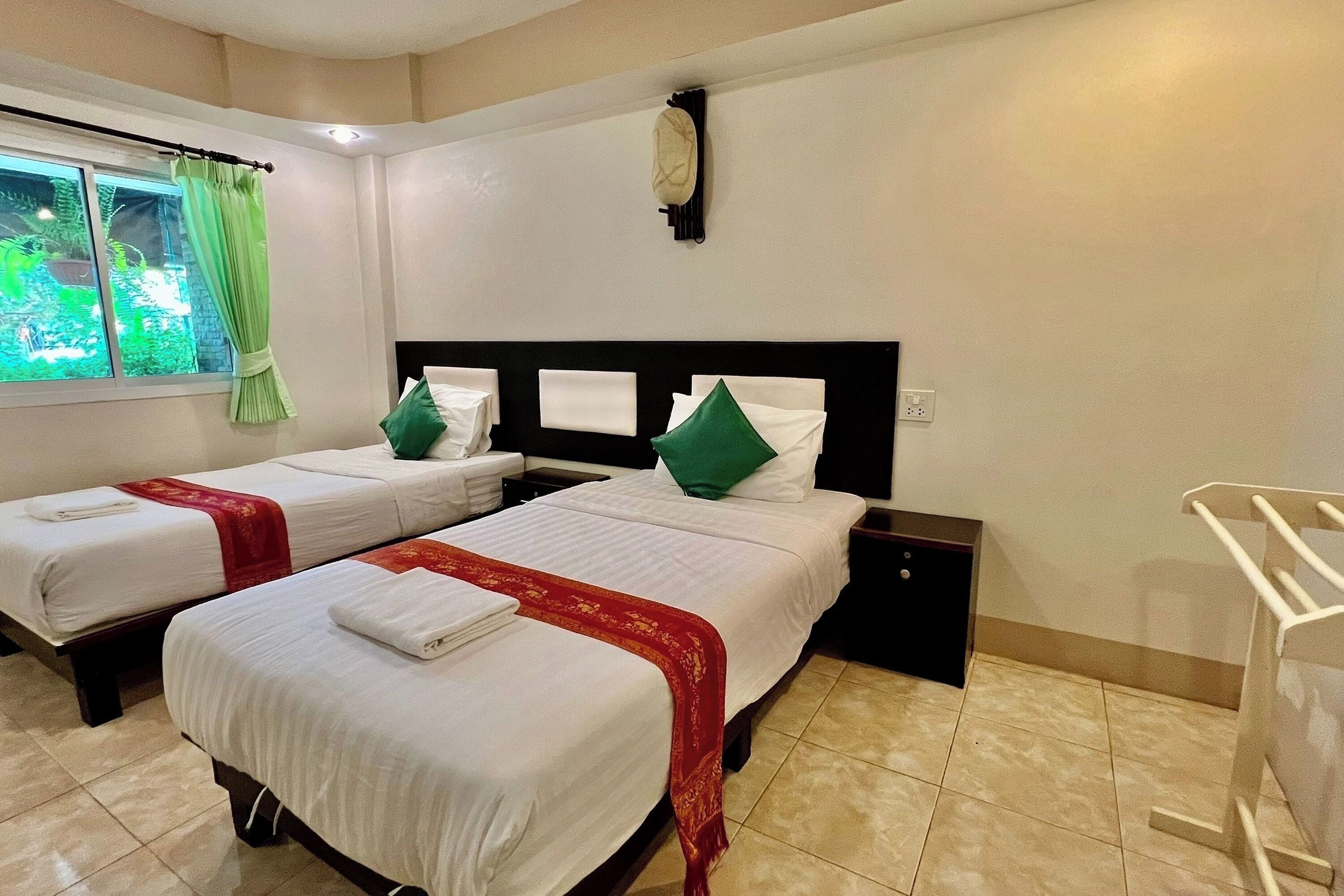 A hotel room with two beds, each with white linens and a red decorative border. There is a window with green curtains, a wall-mounted clock, and a white towel rack.