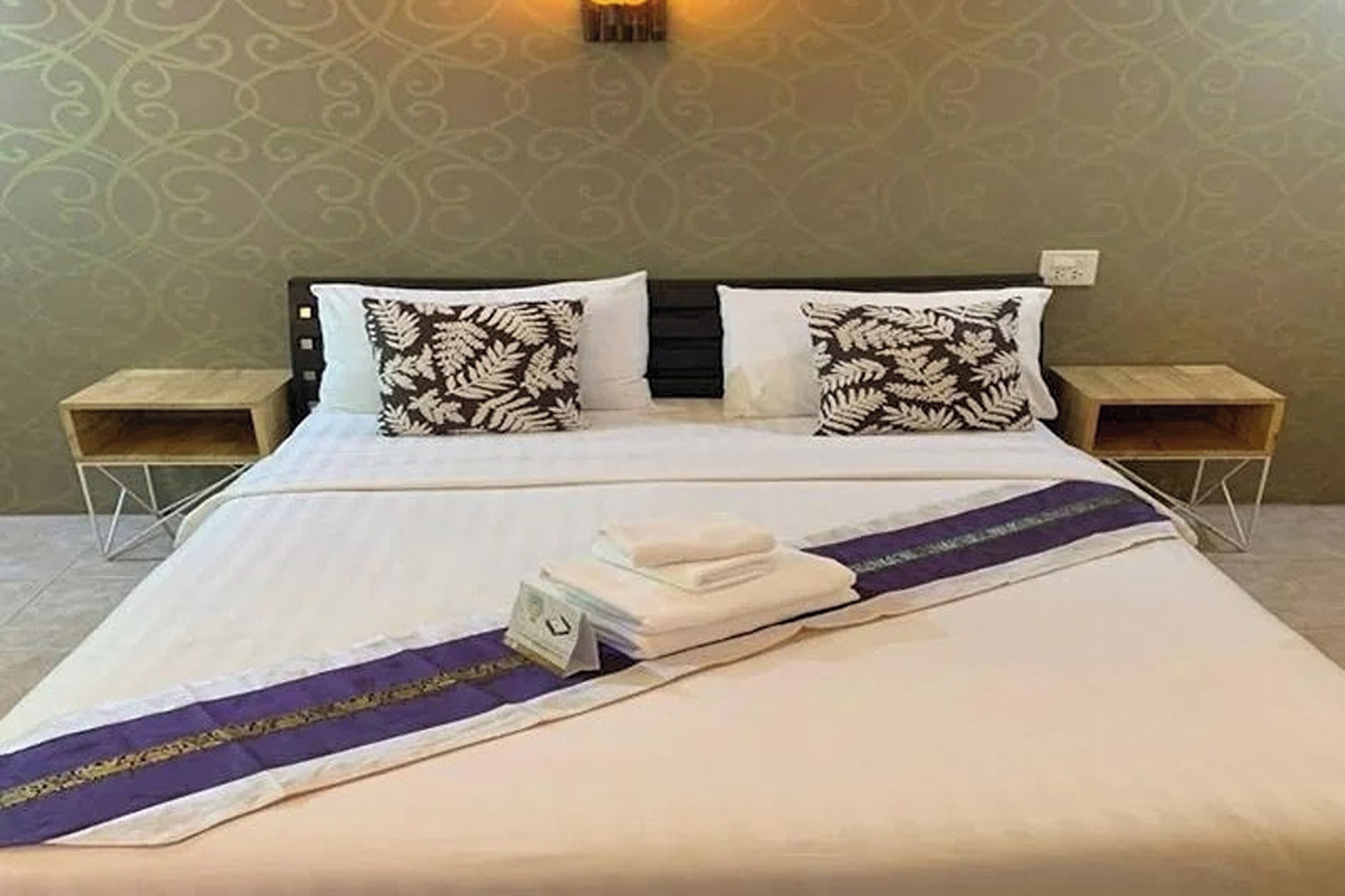 Un lit bien fait, recouvert d’une couverture violette et blanche, avec deux oreillers ornés d’un motif de feuilles et une pile de serviettes pliées posée sur le lit.