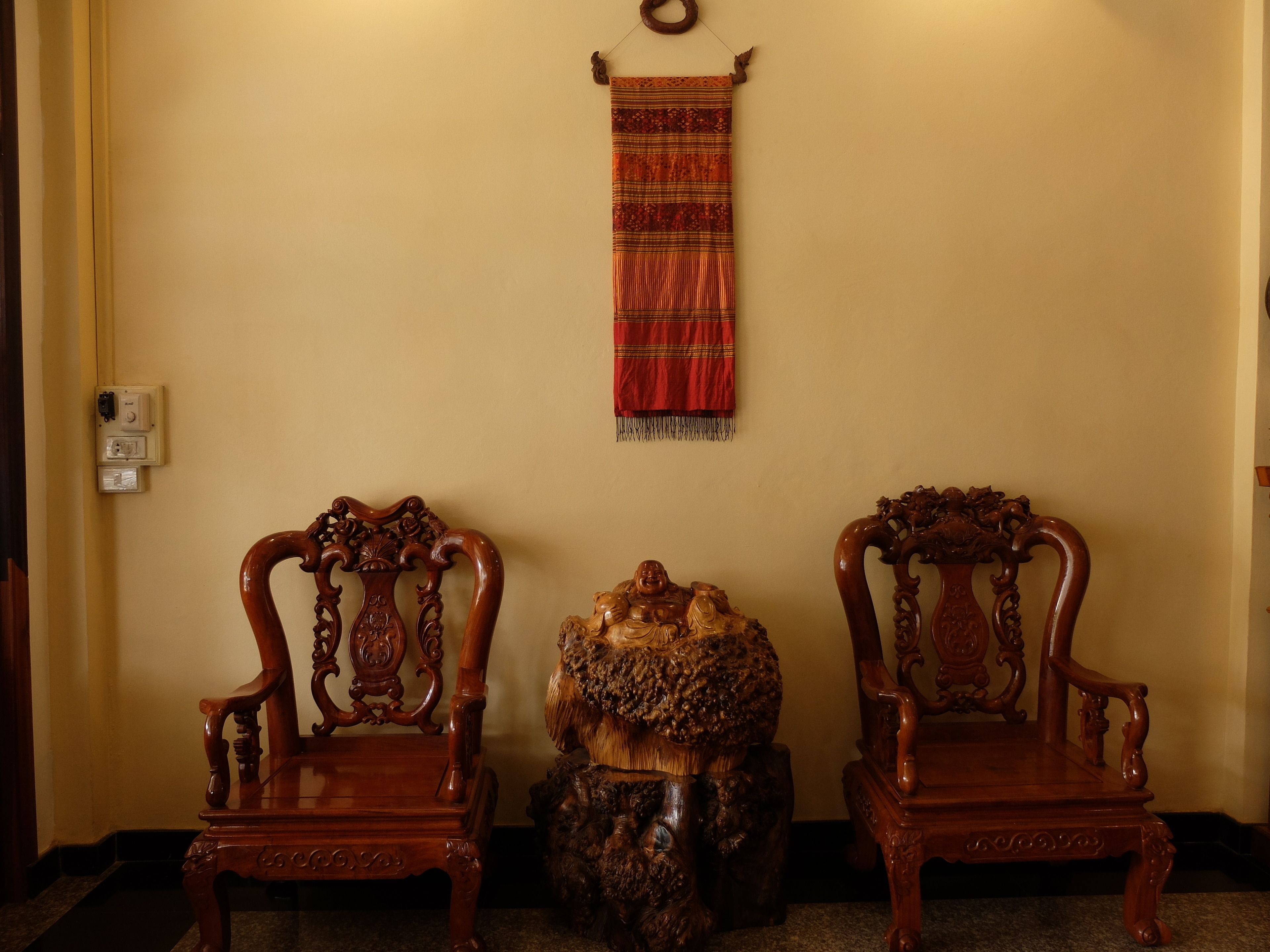 Two ornate wooden chairs with intricate carvings, a decorative wooden sculpture, and a red patterned tapestry hanging on the wall.