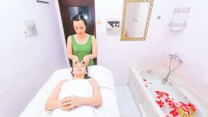 Una persona sdraiata su un letto in una stanza con una vasca da bagno piena di petali di rosa, uno specchio e una piccola radio appesa al muro.