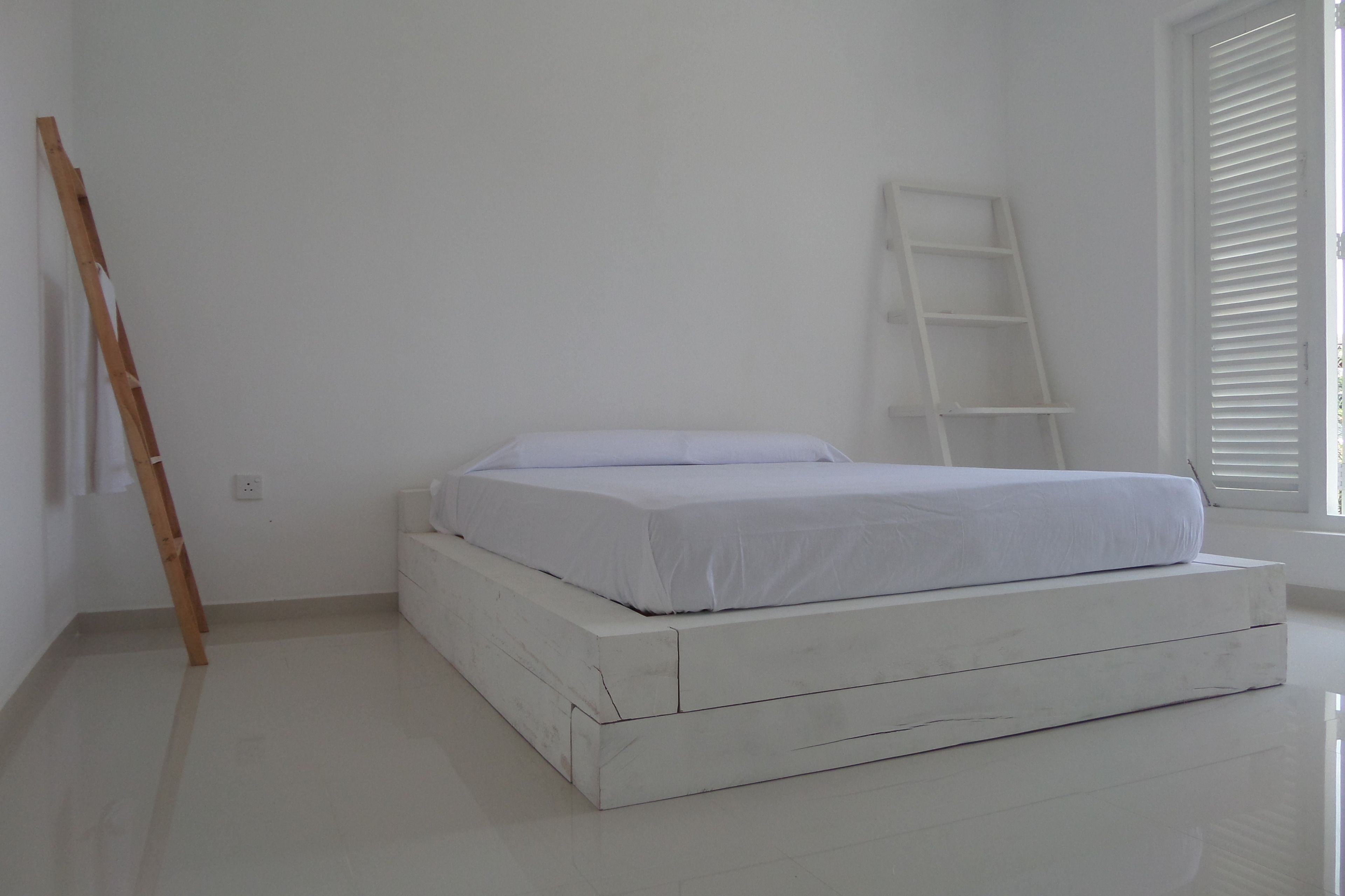 A minimalist room with a white bed, a wooden ladder, and a white shelf unit.