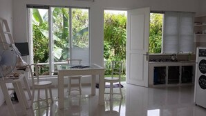 A bright, white room with a table and chairs, a small kitchen area, and large windows with a view of greenery.