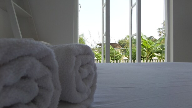 A neatly made bed with white linens and a view of a white fence and greenery outside.