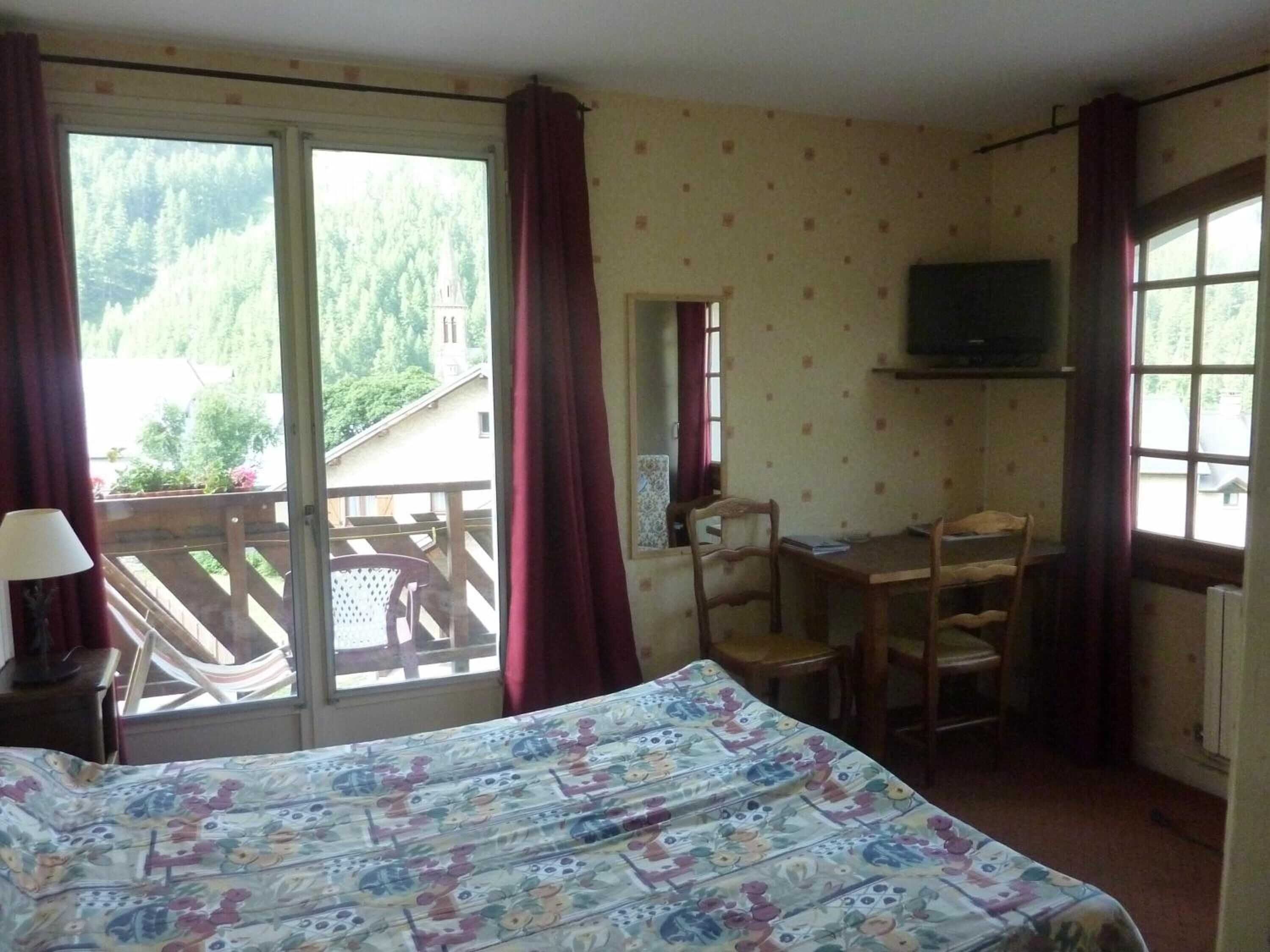 A bedroom with a bed, a desk, and a chair. There is a balcony with a view of trees and a building.