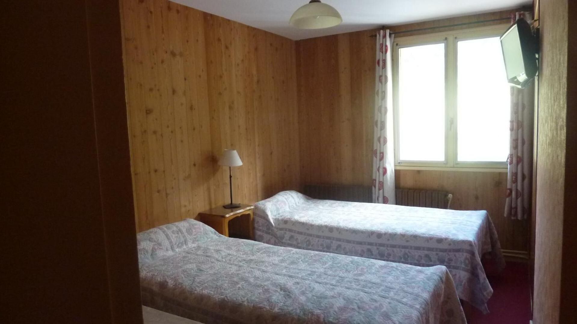 A room with two beds, wooden walls, a window with curtains, a lamp on a nightstand, and a TV mounted on the wall.