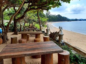 Una mesa de madera y bancos en un espacio al aire libre sombreado, con una playa arenosa y estatuas decorativas al fondo.